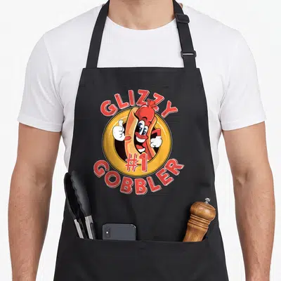 Gobbler hot dog apron - Cartoon hot dog giving thumbs up inside yellow circle with GLIZZY #1 GOBBLER text — shown on a black cotton canvas apron against a white background
