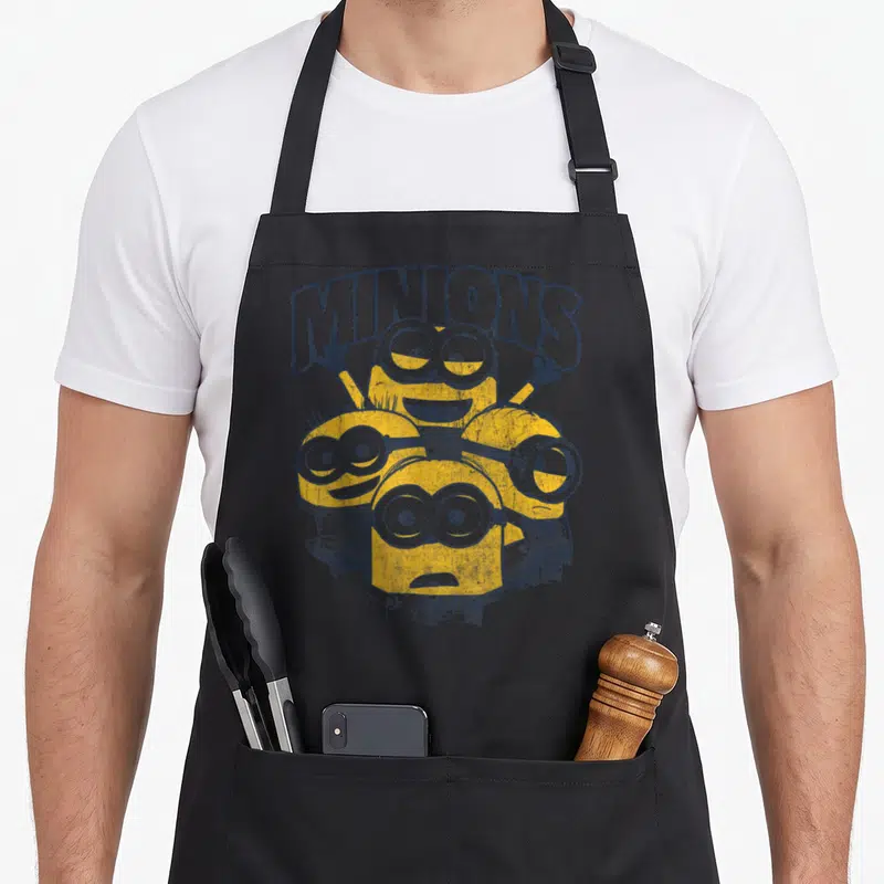 Funny despicable novelty apron - Four yellow minions clustered below arched MINIONS text in dark blue cartoon style — shown on a black cotton canvas apron against a white background