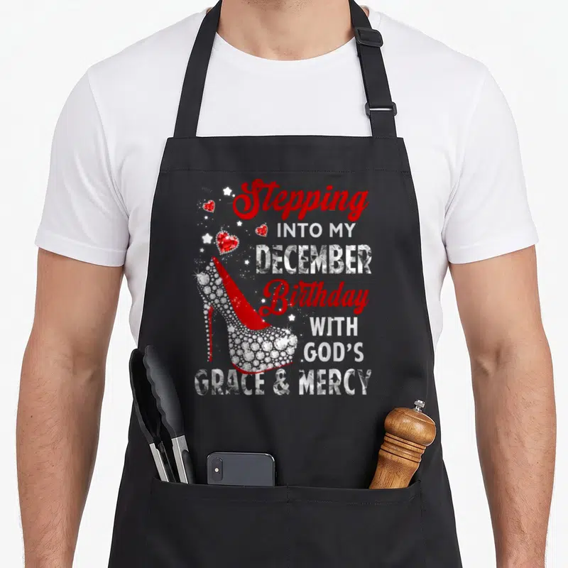 Stepping december birthday apron - Red jeweled high heel shoe beside stacked text Stepping Into My December Birthday — shown on a black cotton canvas apron against a white background
