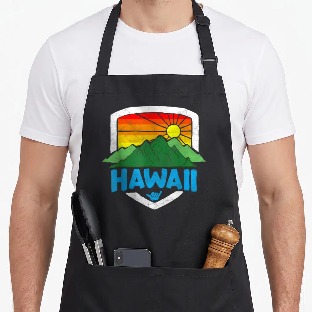 Sun rainbow apron - Retro Hawaii shield with green mountains, striped sunset, blue text, and shaka sign — shown on a black cotton canvas apron against a white background