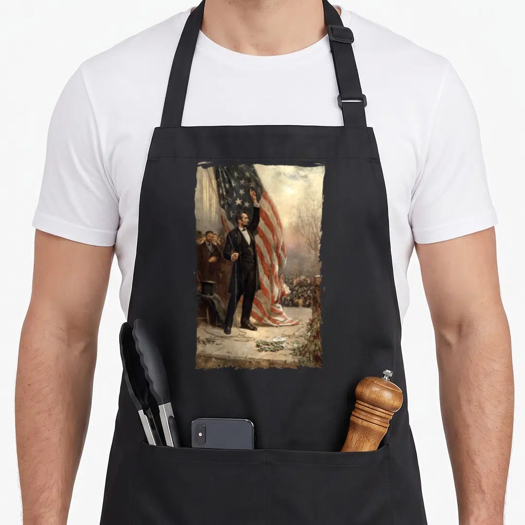 Independence abraham 4th of july apron - Abraham Lincoln raising arm before large draped USA flag with crowd and top — shown on a black cotton canvas apron against a white background