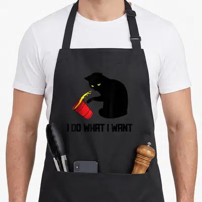 Funny cup cat apron - Black cat silhouette knocking over red cup with yellow spill and text I DO WHAT I WANT — shown on a black cotton canvas apron against a white background