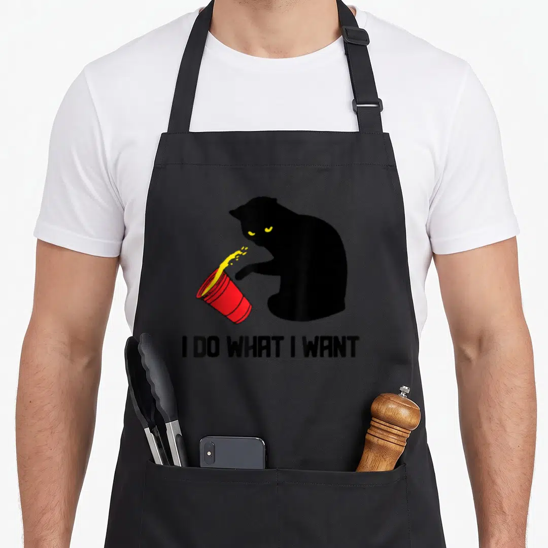 Funny cup cat apron - Black cat silhouette knocking over red cup with yellow spill and text I DO WHAT I WANT — shown on a black cotton canvas apron against a white background