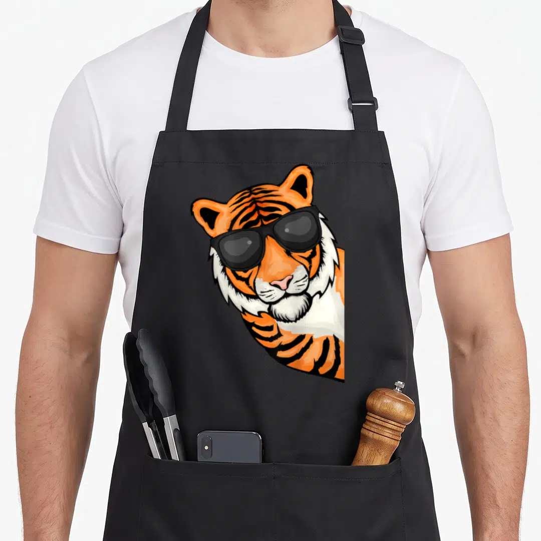 Wildcat tiger apron - Cartoon tiger head wearing black sunglasses in orange and white — shown on a black cotton canvas apron against a white background