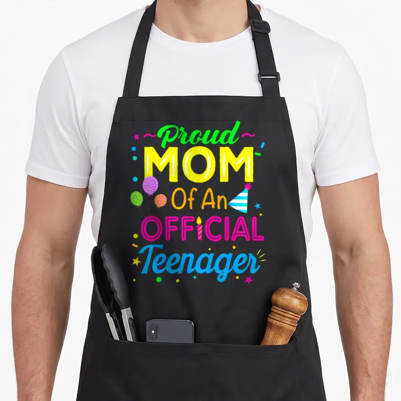 13th birthday apron for the kitchen - Stacked colorful text Proud MOM Of An OFFICIAL Teenager with balloons and party — shown on a black cotton canvas apron against a white background