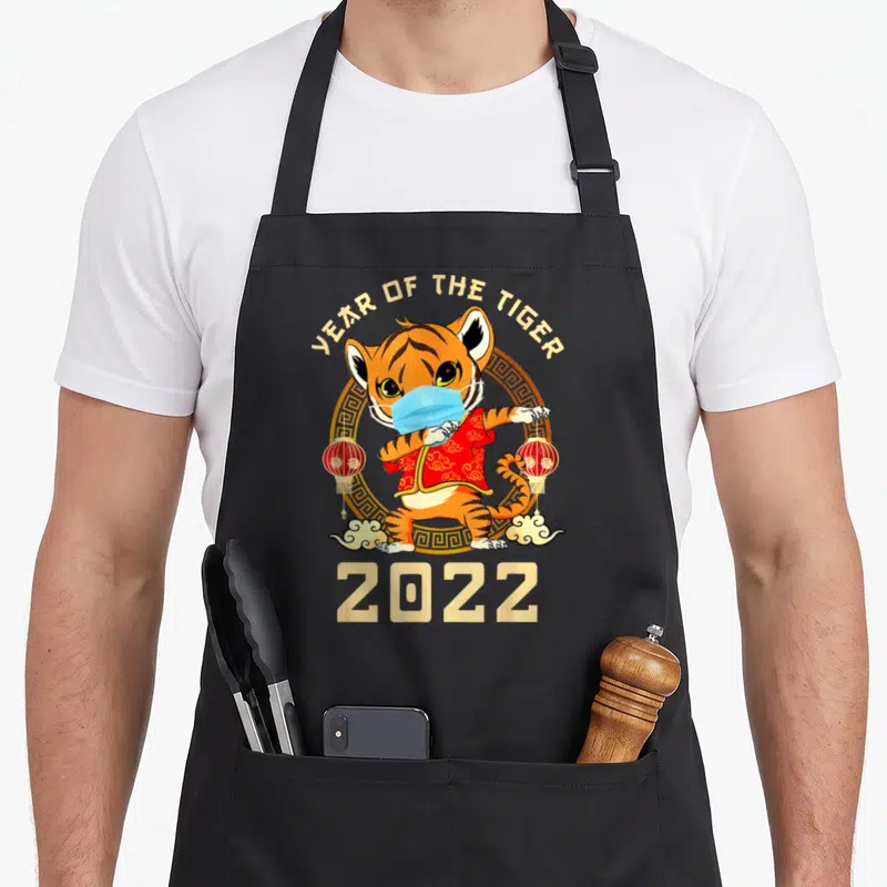 Chinese tiger apron - Cartoon tiger dabbing in a blue mask and red jacket under Year of the Tiger text — shown on a black cotton canvas apron against a white background