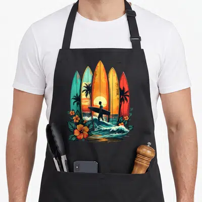 Surfboarder tropical beach apron - Five colorful surfboards frame a surfer silhouette among waves and hibiscus flowers — shown on a black cotton canvas apron against a white background