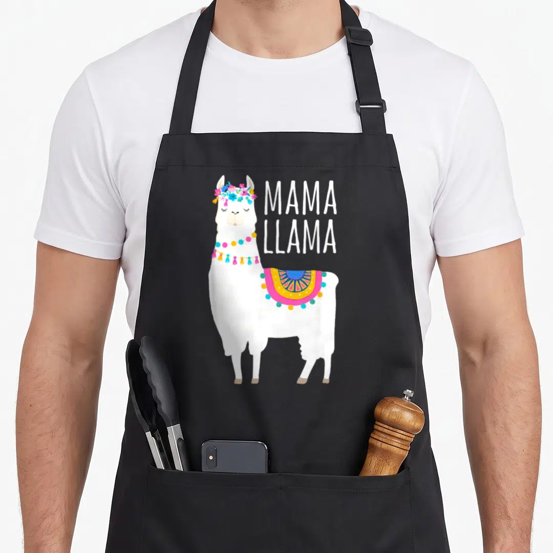 Llama mama apron - White cartoon llama with flower crown, tassel garland, pink blue yellow blanket, and stacked MAMA — shown on a black cotton canvas apron against a white background