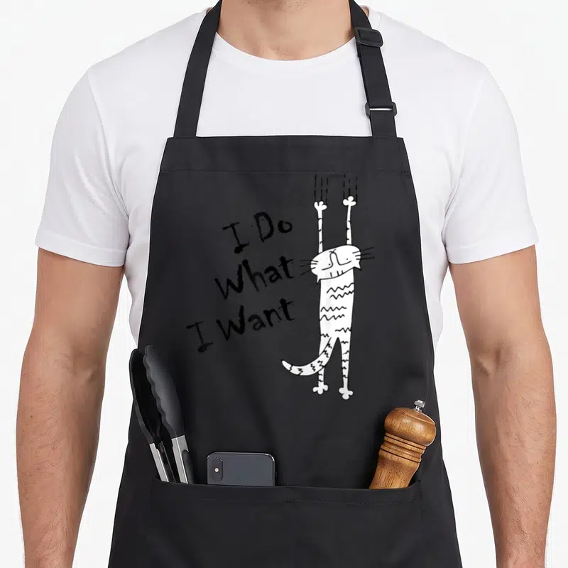 Cute cat apron for baking - White line-art cat hanging vertically next to stacked I Do What I Want text — shown on a black cotton canvas apron against a white background