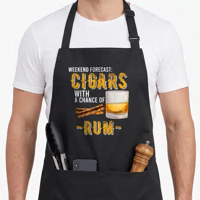Weekend cigar lovers apron - Stacked text Cigars with Chance of Rum next to glass and two brown cigars lying — shown on a black cotton canvas apron against a white background