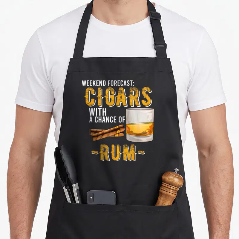 Weekend cigar lovers apron - Stacked text Cigars with Chance of Rum next to glass and two brown cigars lying — shown on a black cotton canvas apron against a white background