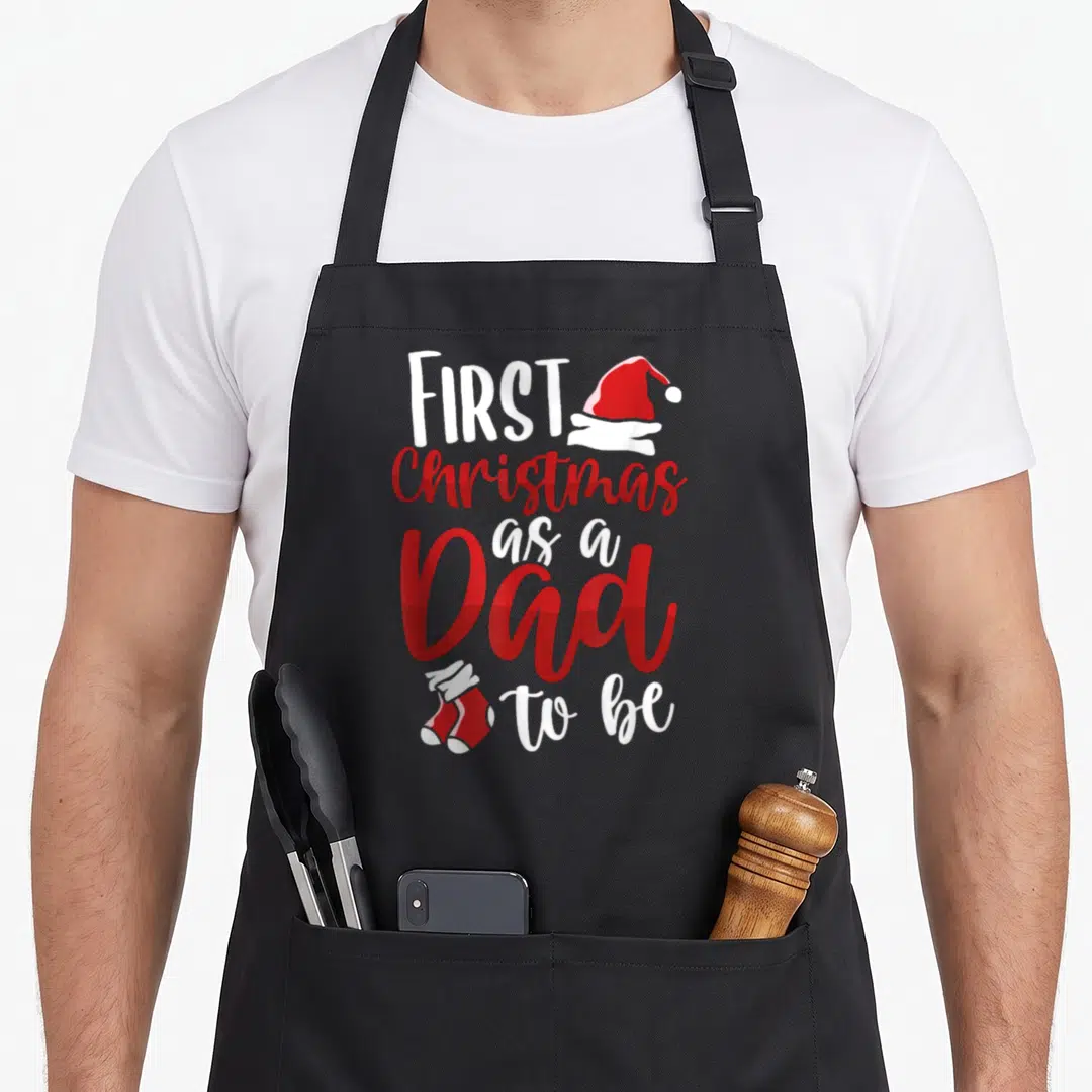 Future christmas aprons - Red Santa hat and two stockings beside stacked white and red text reading FIRST Christmas as — shown on a black cotton canvas apron against a white background