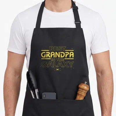 Galaxy father day apron - Yellow BEST GRANDPA IN THE GALAXY text with two crossed lightsabers and scattered stars — shown on a black cotton canvas apron against a white background