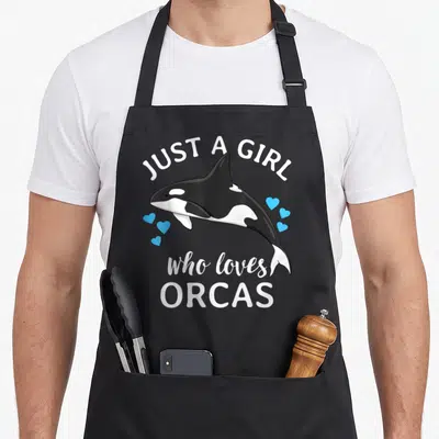 Orcas orca apron - Cartoon orca with white text and scattered blue hearts — shown on a black cotton canvas apron against a white background