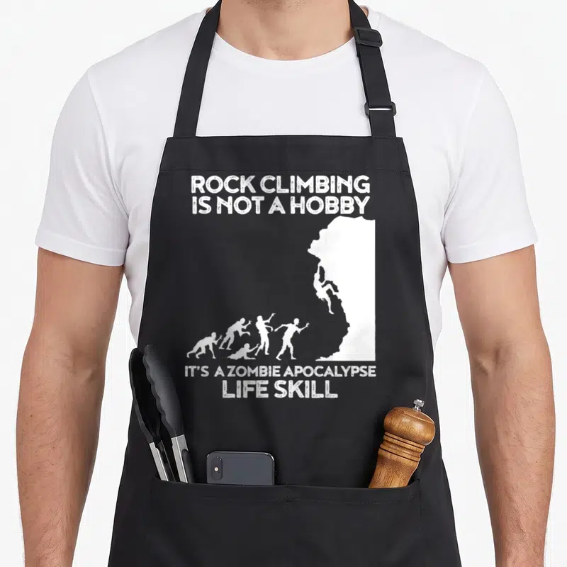 Zombie climbing apron - White text and cliff silhouette with climber and approaching zombies — shown on a black cotton canvas apron against a white background