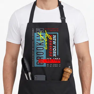 Brooklyn new york apron for men - Vertical BROOKLYN text with yellow scribble, red rectangle over EST 1904 — shown on a black cotton canvas apron against a white background