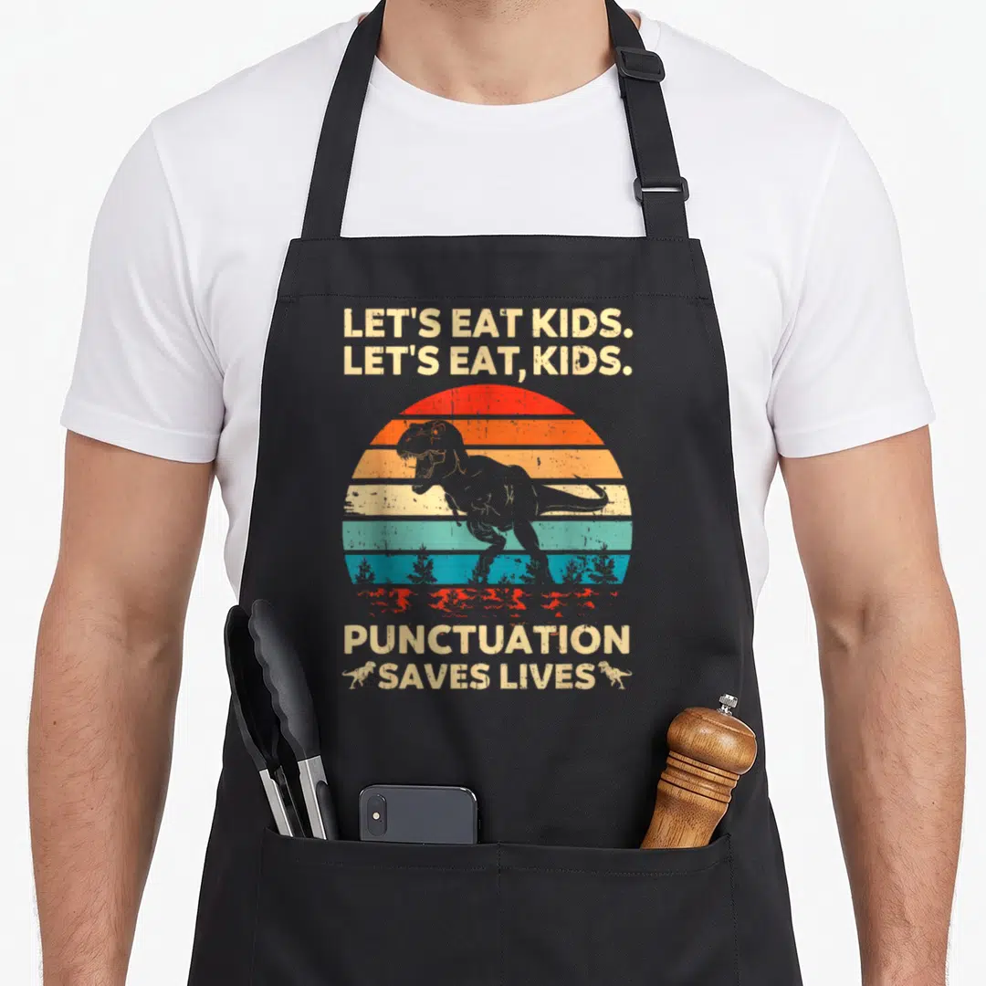 Grammar let's eat apron - Retro T-Rex with striped sun and text Let's Eat Kids Punctuation Saves Lives — shown on a black cotton canvas apron against a white background