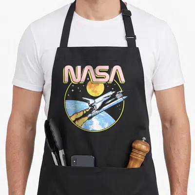 Earth nasa apron - Retro NASA text above shuttle flying over earth moon and stars in pink yellow blue black white — shown on a black cotton canvas apron against a white background