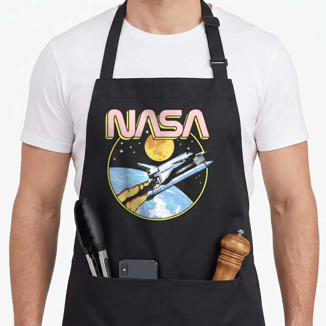 Earth nasa apron - Retro NASA text above shuttle flying over earth moon and stars in pink yellow blue black white — shown on a black cotton canvas apron against a white background