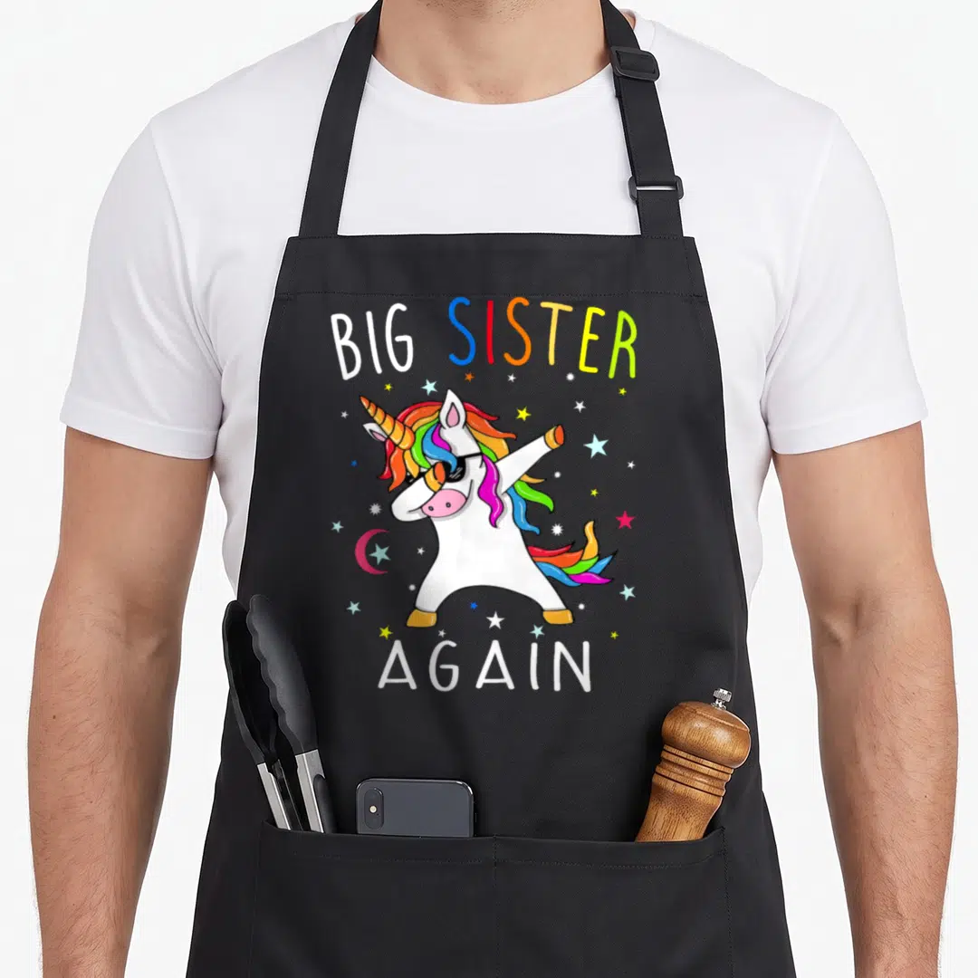 Sibling neat apron - Dabbing unicorn with sunglasses and rainbow mane surrounded by stars and Big Sister Again text — shown on a black cotton canvas apron against a white background