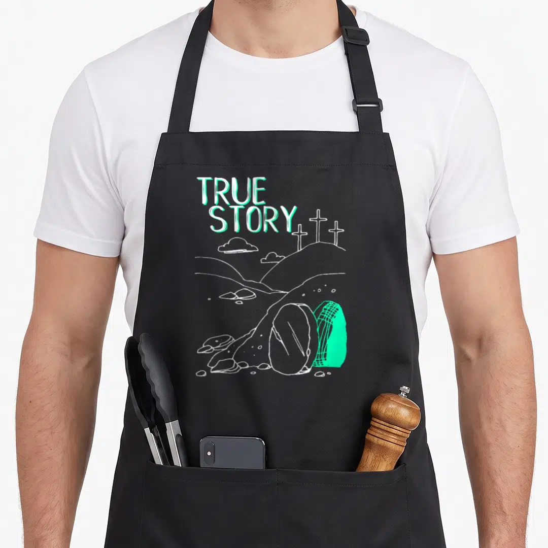 Story easter apron - TRUE STORY text above three crosses and a grave containing a pill and capsule in mint and white — shown on a black cotton canvas apron against a white background