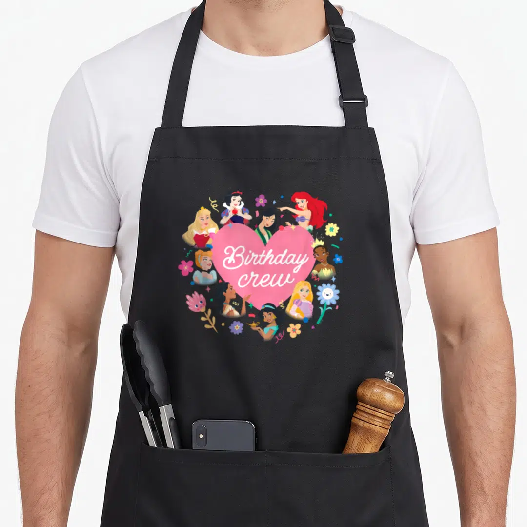 Crew princess apron - Pink heart with Birthday Crew text surrounded by cartoon figures and flowers — shown on a black cotton canvas apron against a white background