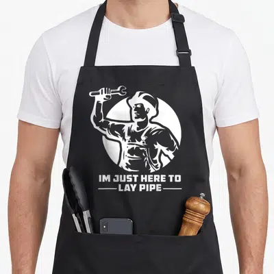 Lay pipe apron - White silhouette of muscular man holding wrench inside circle with text IM JUST HERE TO LAY PIPE — shown on a black cotton canvas apron against a white background