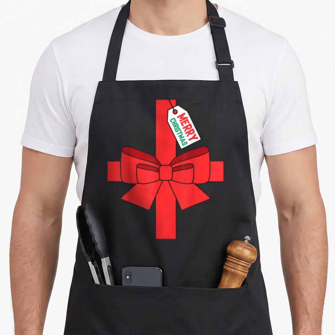 Theme merry christmas apron - Red ribbon cross with large bow and white tag reading MERRY CHRISTMAS in red and green — shown on a black cotton canvas apron against a white background