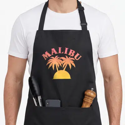 Malibu graphic club inc apron - Pink MALIBU text arching over two orange palm trees and a yellow sun semicircle — shown on a black cotton canvas apron against a white background