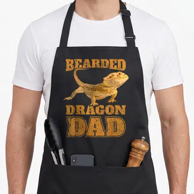 Papa dragon apron - Realistic orange bearded dragon illustration with bold distressed text — shown on a black cotton canvas apron against a white background
