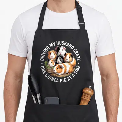 Cavy driving apron - Five cartoon guinea pigs with text driving husband crazy one guinea pig at a time — shown on a black cotton canvas apron against a white background