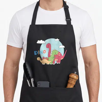Dino road apron - Three cartoon dinosaurs pink green yellow in front of blue circle with DINO text and white clouds — shown on a black cotton canvas apron against a white background
