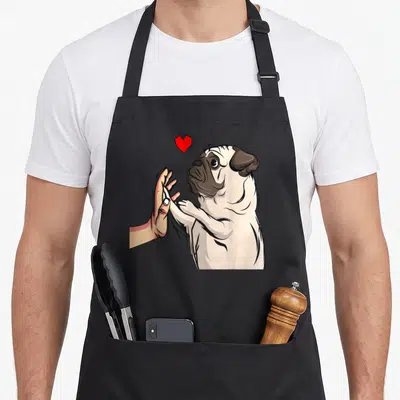 Pug mummy and me aprons - Cartoon pug high-fiving human hand with red heart floating above head — shown on a black cotton canvas apron against a white background