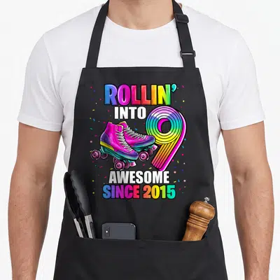 Skating 9th birthday apron - Two pink roller skates over rainbow number 9 with ROLLIN INTO AWESOME SINCE 2015 text — shown on a black cotton canvas apron against a white background
