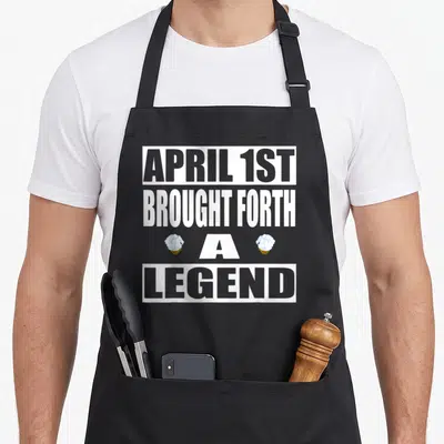 Birthstone aries kitchen apron - APRIL 1ST BROUGHT FORTH A LEGEND text with two blue cupcakes flanking letter — shown on a black cotton canvas apron against a white background