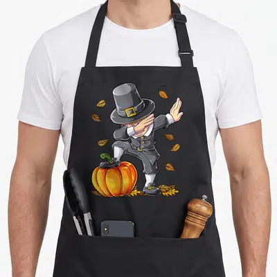Dabbing pilgrim thanksgiving day aprons - Cartoon pilgrim dabbing on pumpkin with scattered autumn leaves in grey — shown on a black cotton canvas apron against a white background
