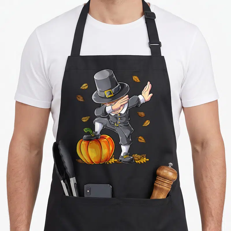 Dabbing pilgrim thanksgiving day aprons - Cartoon pilgrim dabbing on pumpkin with scattered autumn leaves in grey — shown on a black cotton canvas apron against a white background