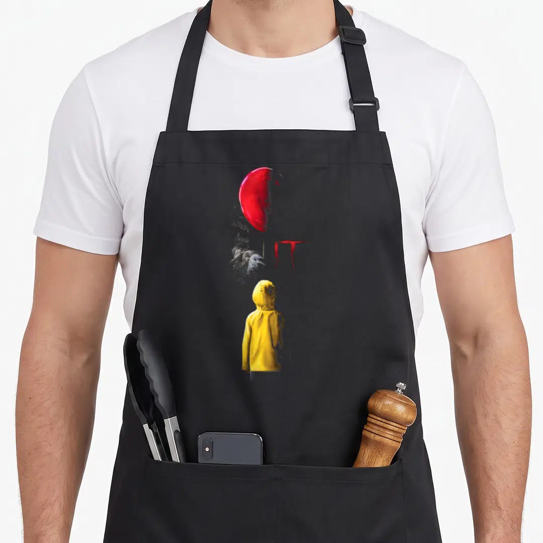 Funny balloon apron - Red balloon above yellow raincoat child facing grey sketch of Pennywise hand and IT text — shown on a black cotton canvas apron against a white background