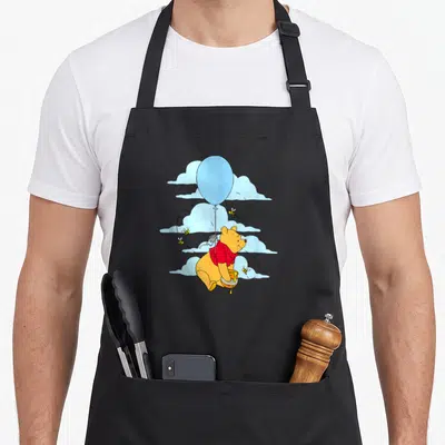 Floating balloon apron - Cartoon Winnie the Pooh in red shirt holding honey pot under blue balloon with bees and clouds — shown on a black cotton canvas apron against a white background