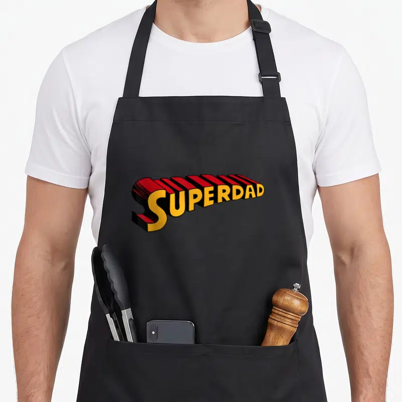 Superdad superhero apron - 3D SUPERDAD text in yellow red and black block letters — shown on a black cotton canvas apron against a white background