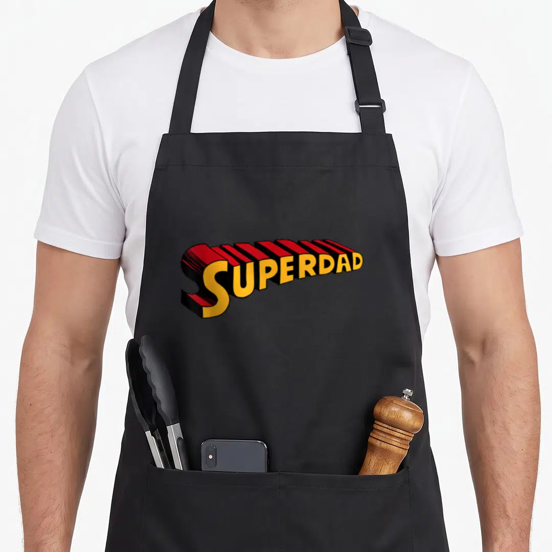 Superdad superhero apron - 3D SUPERDAD text in yellow red and black block letters — shown on a black cotton canvas apron against a white background