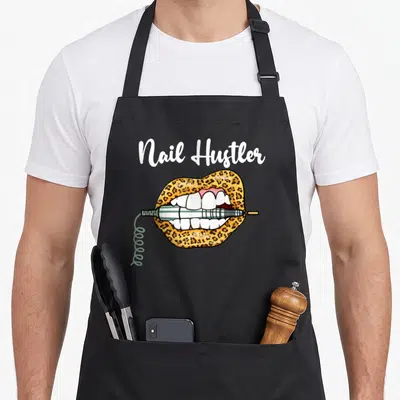 Lip leopard apron - Nail Hustler text above leopard print lips biting a blue electric drill with coiled cord — shown on a black cotton canvas apron against a white background