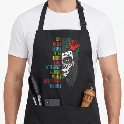 Soy sugar skull apron - Stacked colorful Soy Mujer Latina words beside half sugar skull face with red rose — shown on a black cotton canvas apron against a white background