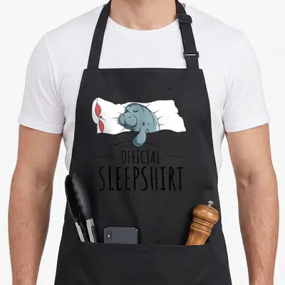 Manatee novelty apron - Cartoon manatee sleeping on white pillow under grey blanket with Official Sleepshirt text below — shown on a black cotton canvas apron against a white background