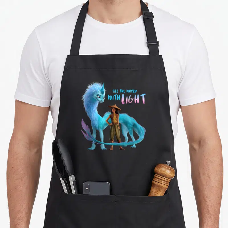 Raya dragon apron - Blue fantasy creature stands behind girl in conical hat with Fill the World with Light text — shown on a black cotton canvas apron against a white background