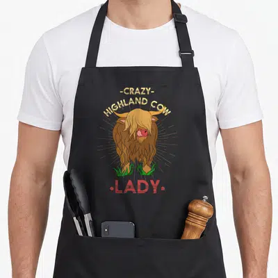 Highland farmer cow print aprons - Cartoon brown highland cow standing in green grass with yellow and red text reading — shown on a black cotton canvas apron against a white background