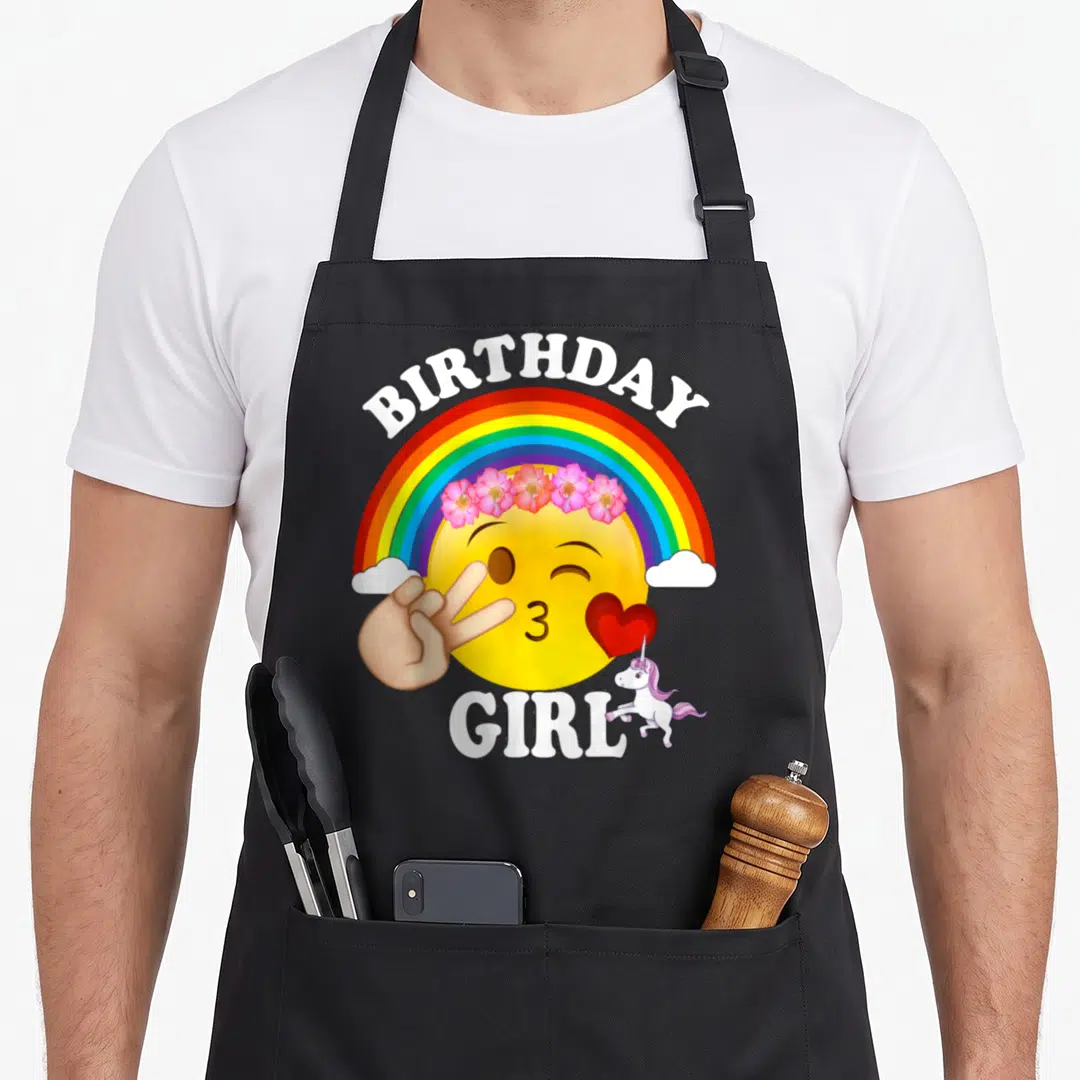 Kiss unicorn apron - Rainbow over yellow emoji with flower crown, peace sign, heart, and unicorn text Birthday Girl — shown on a black cotton canvas apron against a white background
