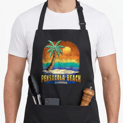 Beach distressed florida apron - Retro palm tree sunset design with Pensacola Beach text and blue ribbon — shown on a black cotton canvas apron against a white background