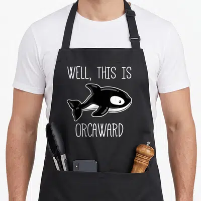 Killer orca lover apron - Cartoon black and white orca with text WELL THIS IS ORCAWARD — shown on a black cotton canvas apron against a white background