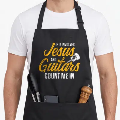 Guitarist christian apron - Orange and white distressed text IF IT INVOLVES Jesus AND Guitars COUNT ME IN with small — shown on a black cotton canvas apron against a white background